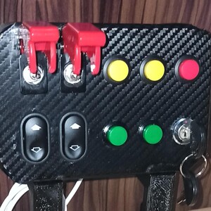 Puede incluir: Un panel negro con un patrón de fibra de carbono, que presenta dos interruptores de palanca rojos, dos interruptores de ventanas, tres botones redondos de color amarillo y verde, y una cerradura con una llave adjunta.
