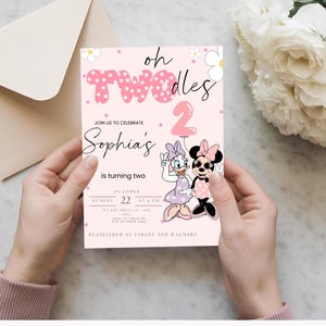 Puede incluir: Una tarjeta de invitación rosa con el texto "oh Twodles" y "Sophia's is turning two". La tarjeta presenta ilustraciones de Minnie y Daisy Mouse, con un globo rosa con el número 2. La invitación está sostenida por manos.