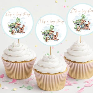 May include: Three cupcakes with white frosting and pink liners. Each cupcake has a round, blue and white paper topper with the text "It's a boy story" and a cartoon image of characters from the Toy Story movie.