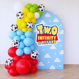 Op de afbeelding: Een kleurrijke ballonnenboog met rode, blauwe, gele, groene en witte ballonnen, sommige versierd met zwarte stippen. Een blauwe boogvormige achtergrond toont witte wolken en de tekst "Two Infinity & Beyond."