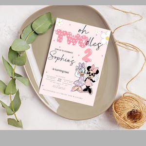 May include: A pink birthday invitation with the text "oh Twodles" and "Sophia's is turning two." Features Daisy Duck and Minnie Mouse. The invitation is on a grey plate with greenery and twine.