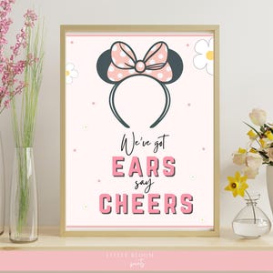 Può includere: Stampa incorniciata con un design di fascia per orecchie di Minnie Mouse e un fiocco rosa a pois. Il testo recita "We've got EARS say CHEERS" in rosa e nero. La stampa è esposta su una mensola con vasi di fiori.