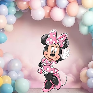Op de afbeelding: Een cartoon illustratie van Minnie Mouse in een roze jurk en strik met stippen, die een vredesteken geeft. De achtergrond toont een roze lucht en een kleurrijke ballonnenboog in pastelkleuren, geschikt voor een verjaardagsfeest of viering.