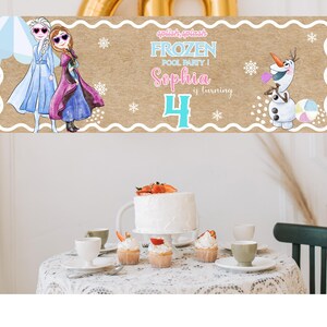 K&ouml;nnte beinhalten: Ein Banner mit dem Text "Frozen Pool Party! Sophia wird 4" mit Illustrationen von Elsa, Anna und Olaf. Das Banner hat einen braunen Hintergrund mit einem wei&szlig;en, gewellten Rand und Schneeflocken-Akzenten. Ein wei&szlig;er Kuchen und Cupcakes stehen auf einem Tisch.