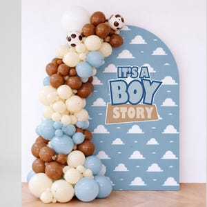 May include: A blue arch-shaped sign with white clouds and the text "IT'S A BOY STORY" is decorated with a balloon garland. The garland features white, brown, and blue balloons, with soccer ball balloons at the top.