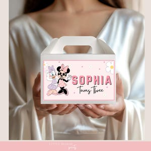 May include: A white party favor box with a pink label featuring Minnie Mouse and Daisy Duck. The label reads "SOPHIA Turns Three" in pink lettering, with daisy and dot accents. The box has a handle.