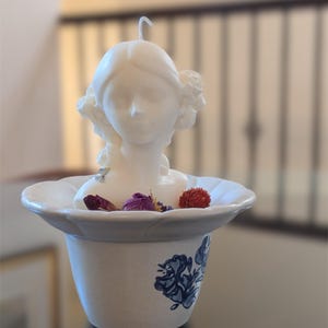 Puede incluir: Una vela de cera blanca con forma de busto humano y una mecha central, colocada dentro de un cuenco de cerámica claro con un patrón floral azul. El cuenco contiene pequeños capullos de flores secas rojas y moradas alrededor de la base.