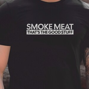 May include: Black crew neck t-shirt featuring the text "SMOKE MEAT" above "THAT'S THE GOOD STUFF" in a white, distressed font. The shirt is a casual style.