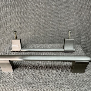 May include: Two brushed metal cabinet pulls. One pull is mounted with screws, the other is lying flat. The pulls have a rectangular base and a curved handle. The hardware is a muted silver color.