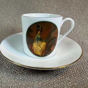 May include: A white ceramic mug with a black handle and the text "But First, Coffee" in a bold, black font. The mug is set against a blurred background, suggesting a cozy setting.