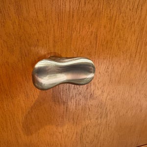 May include: A brushed metal cabinet knob with a unique, elongated shape. The knob is mounted on a wooden cabinet with a visible wood grain pattern. The knob's design is a smooth, ergonomic shape, providing a modern touch to the cabinet.