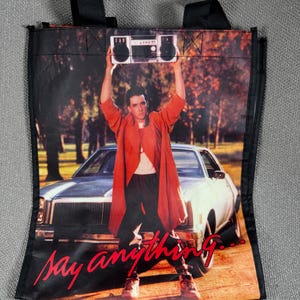 May include: Black tote bag featuring a movie scene with a man holding a boombox above his head in front of a car. The text "Say anything..." is written in red script across the bottom of the bag.