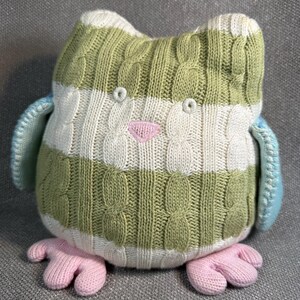 May include: A knitted owl plush toy with green and white striped body, light blue wings, and pink feet. The owl has a pink triangle-shaped beak and small, round eyes. The knitted texture gives the toy a soft, handmade appearance.