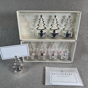 May include: A set of silver Christmas tree place card holders and place cards. The place card holders are in the shape of a Christmas tree. The place cards are white with a black border. The set comes in a clear box.