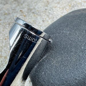 May include: Close-up of a Swatch watch clasp. The clasp is made of shiny, polished metal and features the brand name. The watch clasp is next to a dark gray, textured stone.
