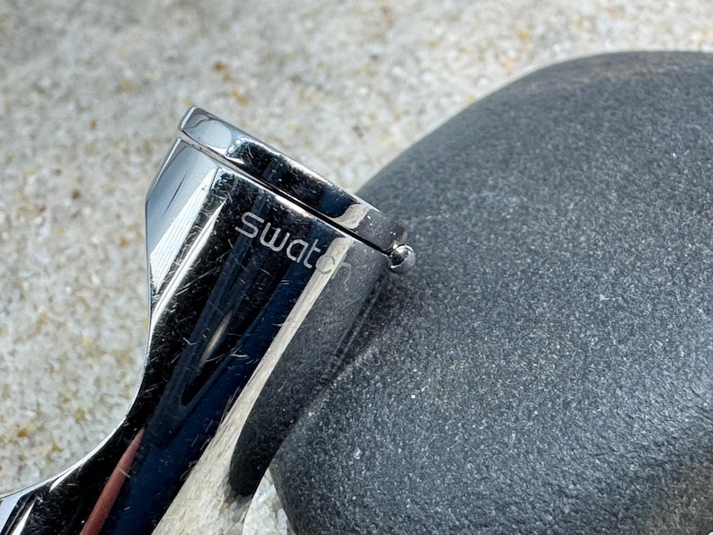 May include: Close-up of a Swatch watch clasp. The clasp is made of shiny, polished metal and features the brand name. The watch clasp is next to a dark gray, textured stone.