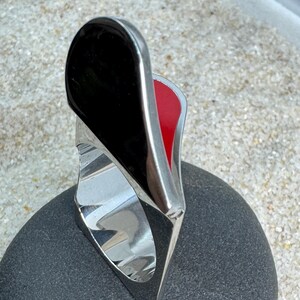 May include: A modern ring with a polished silver band and a striking design. The ring features a large, black, glossy, teardrop-shaped element, with a red interior. The ring is set on a dark gray stone.