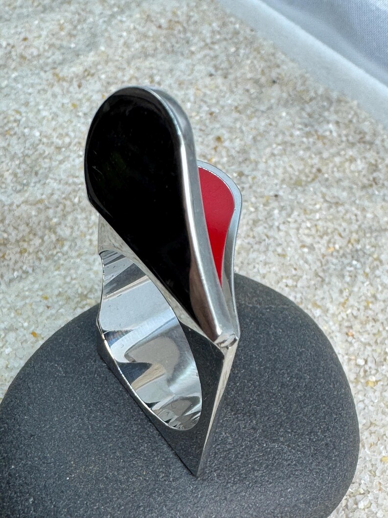 May include: A modern ring with a polished silver band and a striking design. The ring features a large, black, glossy, teardrop-shaped element, with a red interior. The ring is set on a dark gray stone.