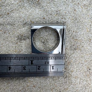 May include: A silver-colored ring with a square design and a circular opening. The ring is measured with a ruler, showing a width of approximately 1 inch. The ring is displayed on a sandy surface.