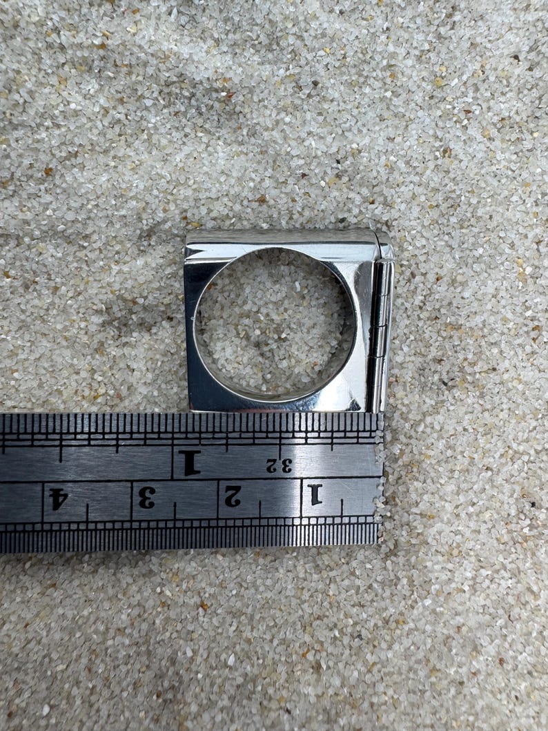 May include: A silver-colored ring with a square design and a circular opening. The ring is measured with a ruler, showing a width of approximately 1 inch. The ring is displayed on a sandy surface.