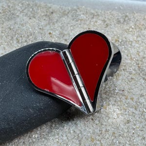 May include: A heart-shaped pendant with a hinged design, featuring a vibrant red enamel filling and a polished silver-tone metal frame. The pendant is resting on a dark gray stone, with a sandy background.