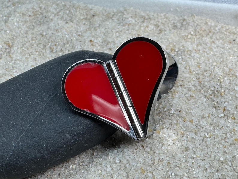 May include: A heart-shaped pendant with a hinged design, featuring a vibrant red enamel filling and a polished silver-tone metal frame. The pendant is resting on a dark gray stone, with a sandy background.