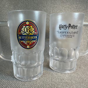 May include: Two frosted glass beer mugs. One mug features a colorful Butterbeer label, the other has "Harry Potter: A Forbidden Forest Experience" printed on it. Both mugs have a handle and a textured base.