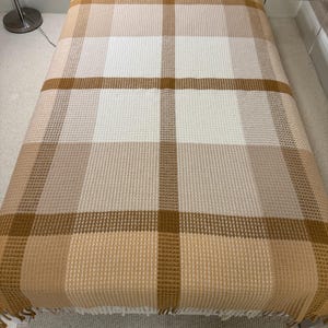 May include: A waffle-weave blanket in a plaid pattern, with tan, cream, and brown hues. The rectangular blanket has a fringed edge. The blanket is laid out on a bed.