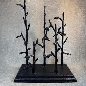 May include: A black metal display stand featuring four vertical branches of varying heights attached to a rectangular base. The structure mimics the appearance of a small forest with thin, textured limbs and small leaf-like protrusions.