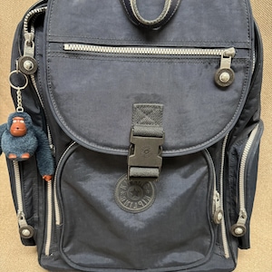 May include: Navy blue backpack with multiple zippered compartments and a flap closure. Features a top handle, adjustable straps, and a small monkey keychain. The backpack has a logo patch on the front and silver-toned hardware.