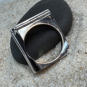 May include: A square, silver-toned ring with a hinged top, resting on a dark gray stone. The ring has a polished finish and a circular opening. The background is a light-colored, sandy texture.