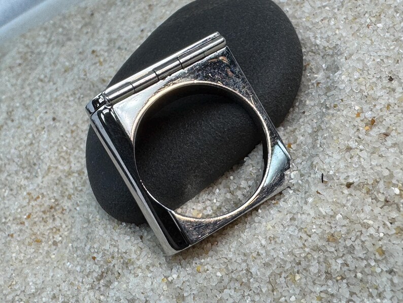 May include: A square, silver-toned ring with a hinged top, resting on a dark gray stone. The ring has a polished finish and a circular opening. The background is a light-colored, sandy texture.