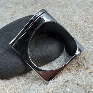 May include: A shiny, silver-toned ring with a square design. The ring is set on a dark gray stone, with a background of white sand. The ring's geometric shape and polished surface create a modern, minimalist aesthetic.