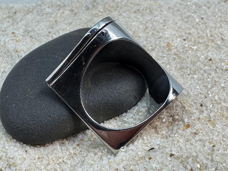May include: A shiny, silver-toned ring with a square design. The ring is set on a dark gray stone, with a background of white sand. The ring's geometric shape and polished surface create a modern, minimalist aesthetic.