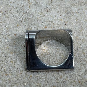 May include: A square, silver-toned ring with a rectangular band and a large, open square center. The ring is set against a background of small, light-colored pebbles. The ring's design is modern and minimalist.
