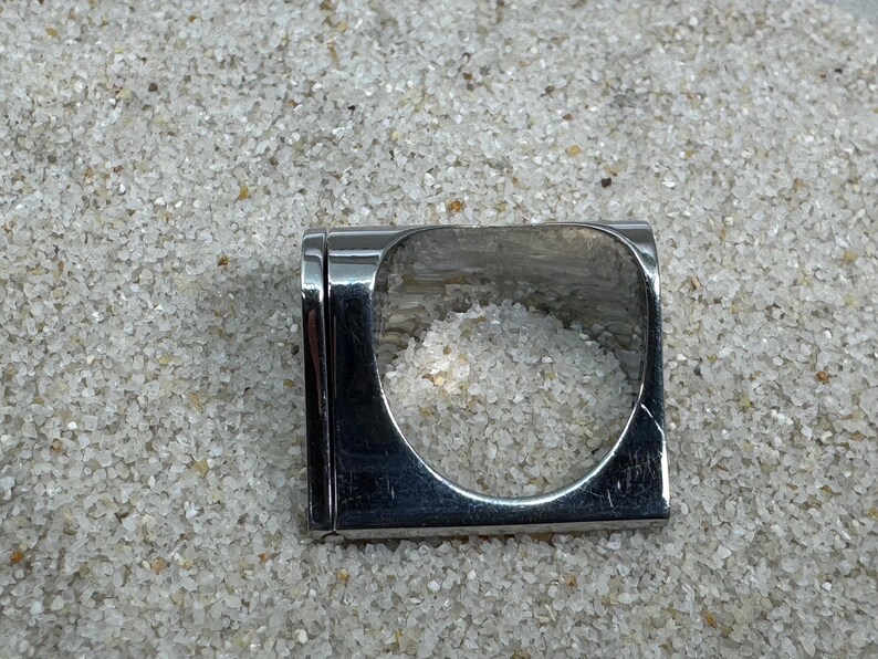 May include: A square, silver-toned ring with a rectangular band and a large, open square center. The ring is set against a background of small, light-colored pebbles. The ring's design is modern and minimalist.