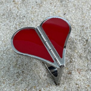 May include: A heart-shaped ring with a hinged design. The ring features a vibrant red enamel heart set within a silver-toned metal frame. The ring is resting on a sandy surface.
