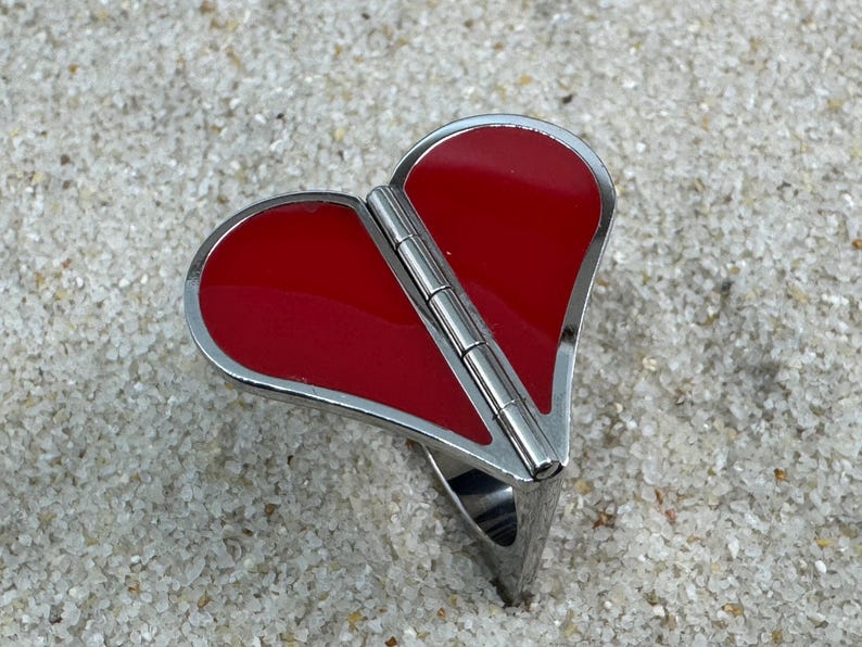 May include: A heart-shaped ring with a hinged design. The ring features a vibrant red enamel heart set within a silver-toned metal frame. The ring is resting on a sandy surface.