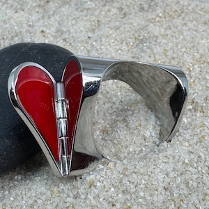 May include: A silver-toned cuff bracelet with a hinged, heart-shaped centerpiece. The heart is filled with glossy red enamel. The bracelet is resting on a dark gray stone and a bed of small, light-colored grains.