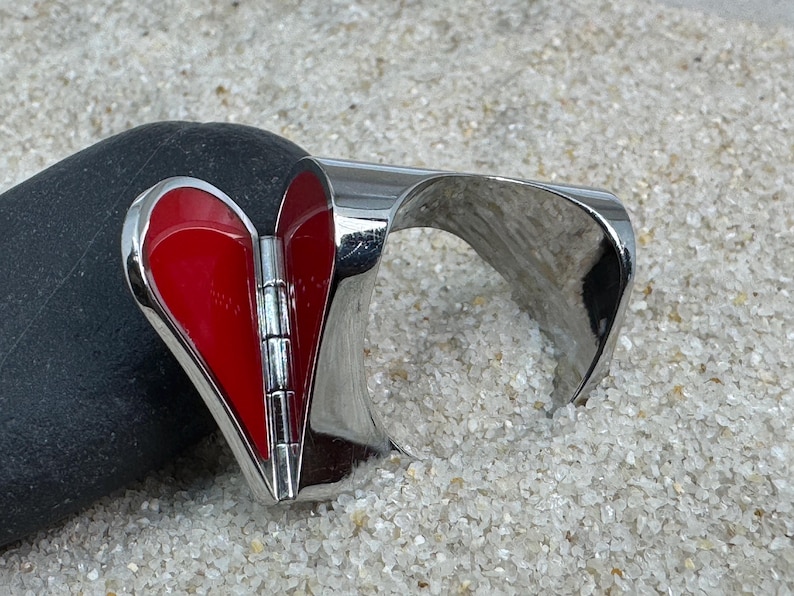 May include: A silver-toned cuff bracelet with a hinged, heart-shaped centerpiece. The heart is filled with glossy red enamel. The bracelet is resting on a dark gray stone and a bed of small, light-colored grains.