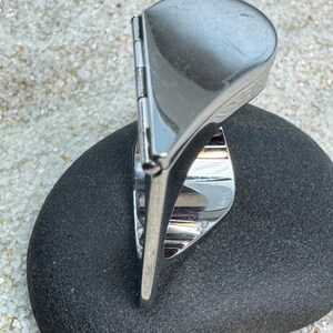 May include: A silver-toned ring with a unique design, featuring a rounded top and a hinged element. The ring is set on a dark gray stone, with a sandy background.