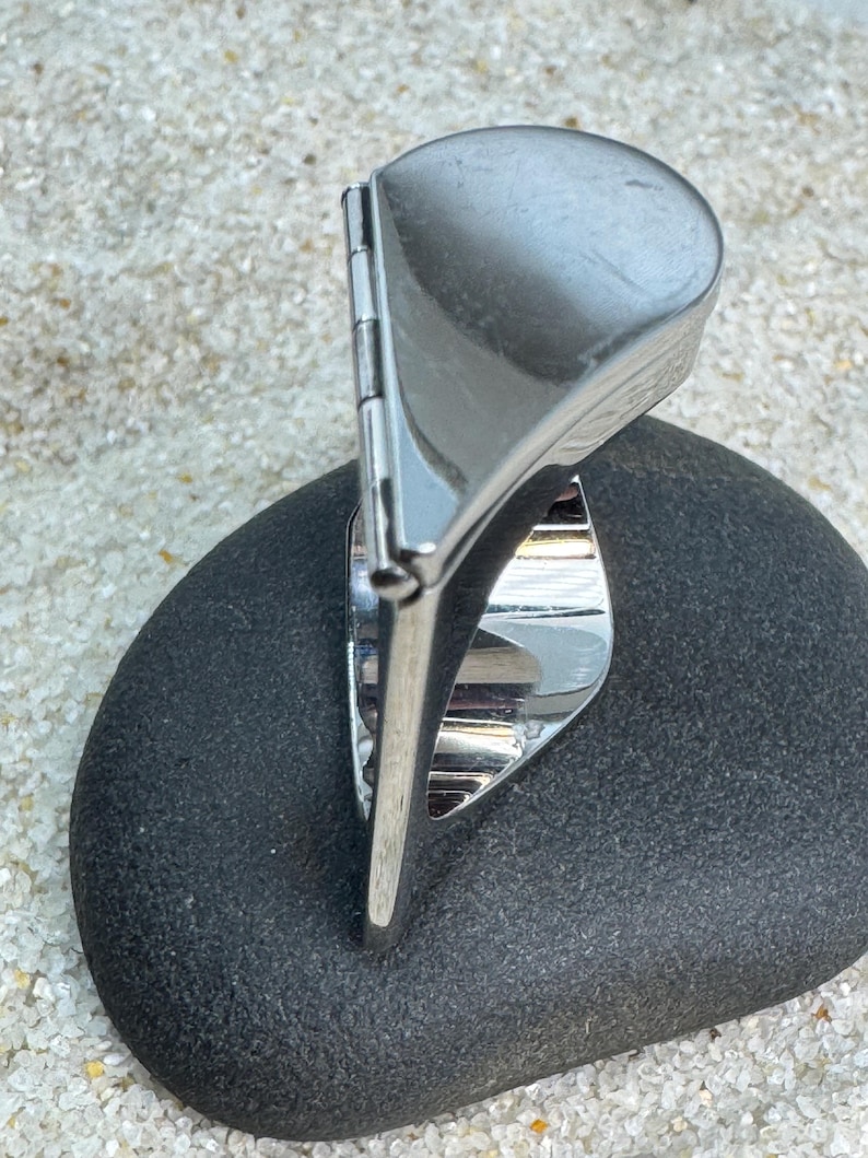 May include: A silver-toned ring with a unique design, featuring a rounded top and a hinged element. The ring is set on a dark gray stone, with a sandy background.