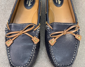 May include: A pair of navy blue leather loafers with tan accents and white stitching. The shoes feature a bow detail and the brand name "Clarks Artisan" is visible on the insole. These shoes are a classic style.