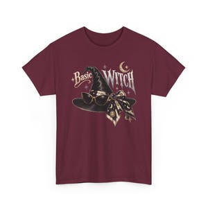 May include: A maroon short-sleeve t-shirt featuring a graphic of a black witch hat adorned with a patterned bow and sunglasses. The text "Basic WITCH" is printed above the hat, accompanied by celestial symbols including a crescent moon and stars.