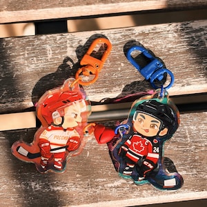 May include: Two hockey-themed keychains. One keychain features a cartoon hockey player in a red and white uniform with the word "Россия" on the jersey. The other keychain has a cartoon hockey player in a red and black uniform with a maple leaf.