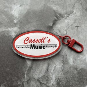 May include: An oval-shaped keyring featuring a white background with a red border. The text 'Cassell's' appears in red script above a black musical staff with notes, followed by the word 'Music' in bold black text. A red metal lobster clasp is attached.