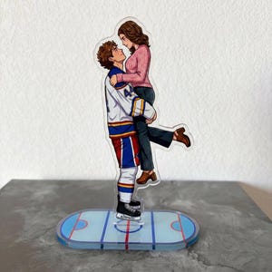 May include: A clear acrylic figurine of a couple embracing. The man, in a hockey jersey, holds the woman. The base is a hockey rink design. The woman wears a pink sweater and the man's jersey has blue, white, and red accents.