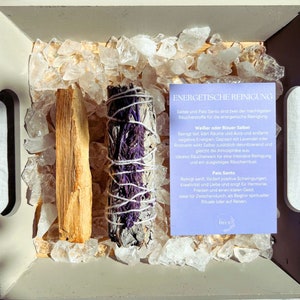 Könnte beinhalten: Nahaufnahme einer Holzkiste, gefüllt mit klaren Quarzkristallen. In der Kiste befinden sich zwei Bündel getrockneter Kräuter, eine weiße Salbei und ein Palo Santo. Eine kleine violette Karte mit dem Text "ENERGETISCHE REINIGUNG" ist in der Kiste sichtbar.