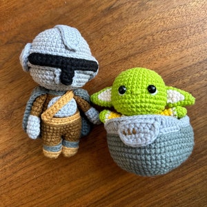 May include: Two crocheted figures: a Mandalorian and Grogu. The Mandalorian is in gray and brown with a black visor. Grogu, in a gray carrier, is green with large black eyes and white-tipped ears. Both are handmade and sit on a wooden surface.