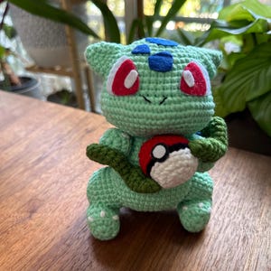 May include: A handmade crochet Bulbasaur plushie in light green, with red and white eyes, blue accents, and a green vine. It holds a red, white, and black Pokeball. The plushie is sitting on a wooden surface.